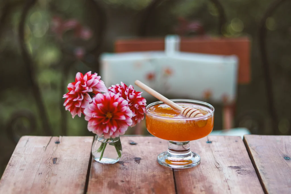 A small vase with pink flowers next to a honey jar on a wooden table outside